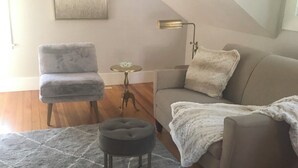 Ein gemütliches Wohnzimmer mit einem grauen Sessel, einer grauen Couch, einem kleinen runden Hocker und einem grauen Teppich. An der Wand hängt ein Gemälde und es steht eine Stehlampe.