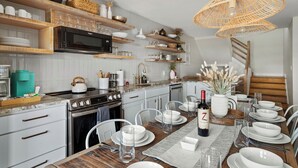A modern kitchen with white cabinetry, wooden shelves, and a dining area set for a meal. - Sea Bound, Private Pool, Fire Pit, Multiple Decks, Hot Tub- 2 Blocks To Beach (Gulf Shores)