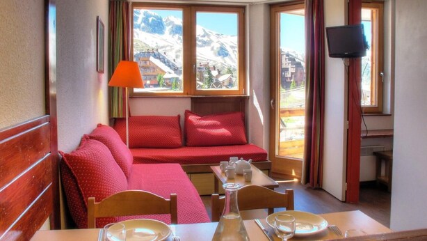 Une pièce chaleureuse avec une table en bois dressée pour le repas, un canapé rouge agrémenté de coussins, une fenêtre donnant sur les montagnes et un téléviseur fixé au mur.