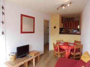 Un espacio habitable compacto con televisor, una mesita, comedor con mesa y sillas, y una cocina con armarios y microondas.
