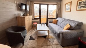 Ein gemütliches Wohnzimmer mit einem grauen Sofa, einem kleinen Tisch, einem dunklen Sessel und einem Holzschrank. Ein Fernseher ist an der Wand befestigt, und es gibt zwei Fenster mit Vorhängen.
