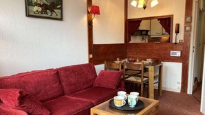 A red sofa, a wooden coffee table with cups, a dining table with chairs, a red lamp, and a painting on the wall.