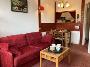 Ein rotes Sofa, ein Couchtisch aus Holz mit Tassen, ein Esstisch mit Stühlen, eine rote Lampe und ein Gemälde an der Wand.