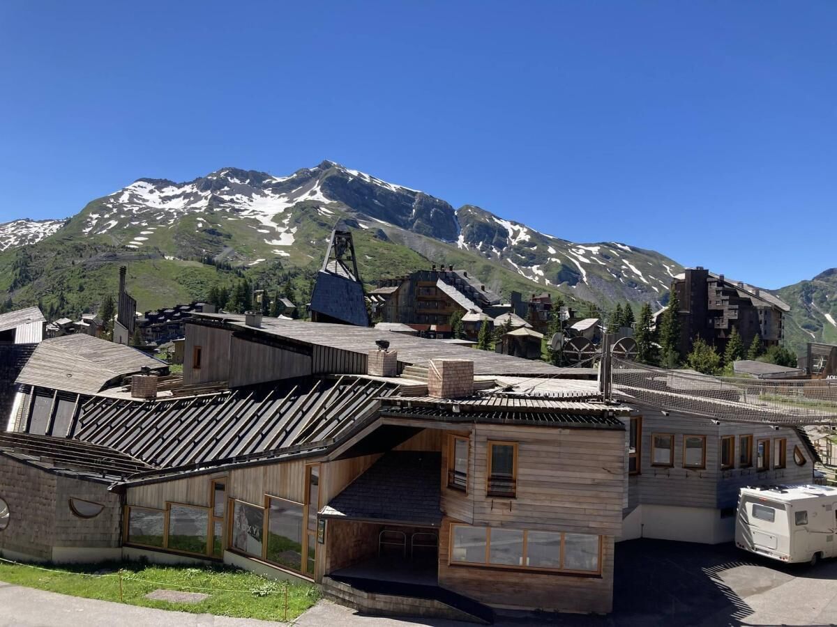 Ein Holzhaus mit geneigtem Dach, einer Treppe, die zu einem Balkon führt, und Blick auf verschneite Berge im Hintergrund.