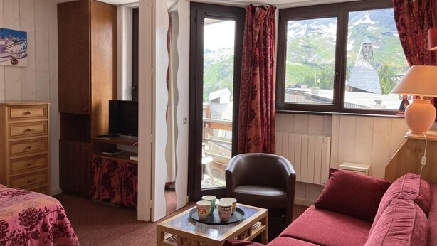 A cozy room with a bed, a sofa, a desk with a chair, and a TV. There is a window with curtains and a view of mountains.