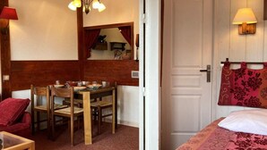 A room with a wooden dining table, chairs, a bed with a floral bedspread, a red pillow, and a mirror above the table.