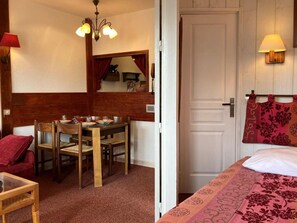 Ein Zimmer mit einem Holztisch, Stühlen, einem Bett mit floralem Überwurf, einem roten Kissen und einem Spiegel über dem Tisch.