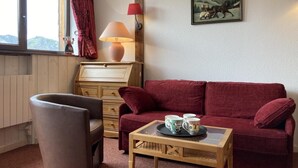 A cozy room with a red sofa, a wooden coffee table with mugs, a wooden cabinet, a lamp, and a window with curtains.