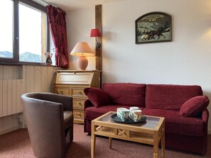 Ein gemütliches Zimmer mit einem roten Sofa, einem Couchtisch aus Holz mit Tassen, einem Holzschrank, einer Lampe und einem Fenster mit Vorhängen.