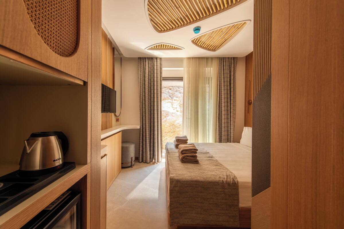 A modern hotel room with a wooden interior, a bed with a beige bedspread, a coffee maker, and a view of the outdoors through the window.