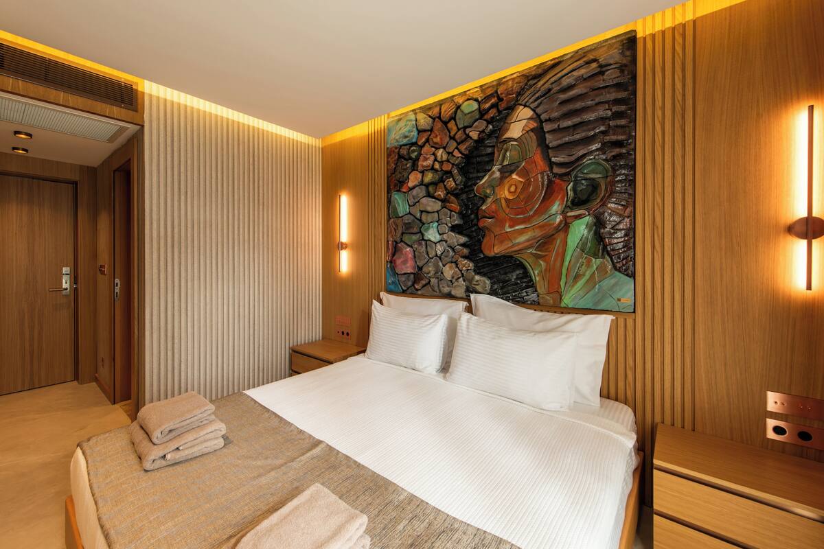 A hotel room with a large bed, a wooden headboard, a stone wall art piece, and a nightstand with a lamp.