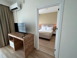 Una habitación de hotel con un escritorio de madera, un televisor de pantalla plana, una cama con ropa blanca y una mesita de noche con una lámpara.