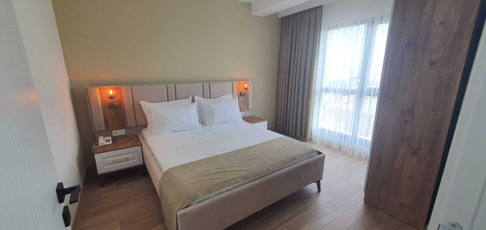 A hotel room with a bed, bedside tables, a nightstand, a window with curtains, and a wooden door.