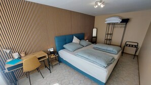 A modern hotel room with a large bed, a desk with a chair, a small table, and a wardrobe.