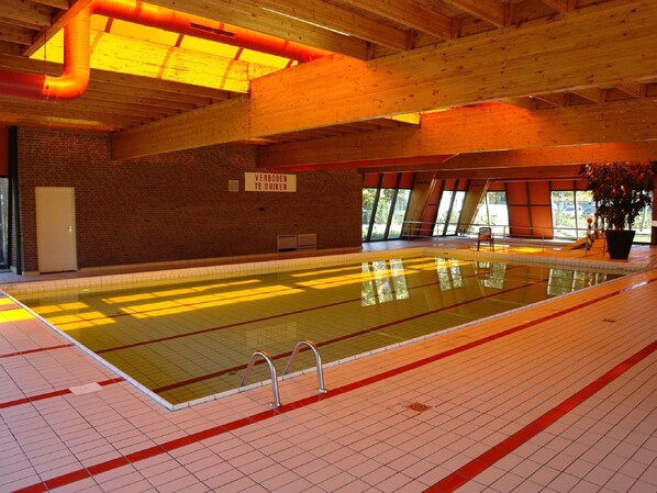 Una piscina interior con suelos y paredes de azulejos, vigas de madera y un cartel en neerlandés.