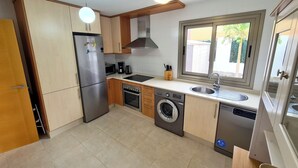 A modern kitchen with wooden cabinets, a stainless steel refrigerator, a built-in oven, a washing machine, and a window providing a view of the outdoors. - Casa Maracuya (Castillo Caleta de Fuste)