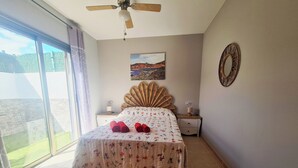 A bedroom with a bed, a ceiling fan, a nightstand, a lamp, a painting, and a window with curtains. - Casa Maracuya (Castillo Caleta de Fuste)