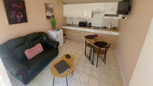 Un espace de vie compact comprenant un canapé, une petite table, un coin cuisine avec des meubles blancs, un four à micro-ondes et un téléviseur fixé au mur.
