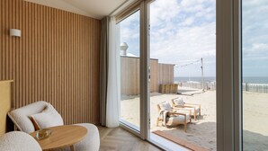 A modern room with a large sliding glass door leading to a beach view. Inside, there is a wooden panel wall, a round wooden table with a glass object, and two beige armchairs. - Beach House in Zandvoort With Sea View (Zandvoort)