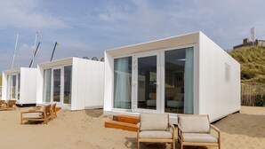 A modern beach house with large sliding glass doors, a wooden deck, and beach chairs. - Beach House in Zandvoort With Sea View (Zandvoort)
