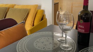 A table with a wine bottle and glasses, a yellow sofa with patterned cushions, and a window with a view of the outside.