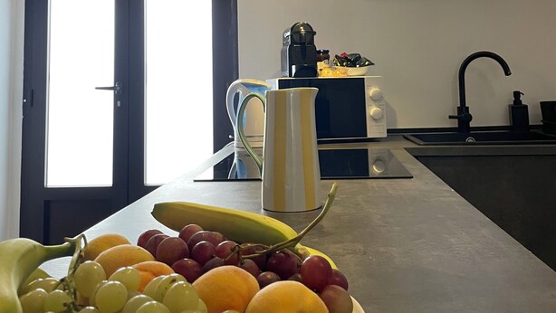 A kitchen with a fruit bowl on the counter, a coffee maker, and a window with a view of the outdoors.