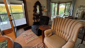 Una sala de estar con un sofá, un sillón y una mesa con objetos decorativos. Hay un ventanal con cortinas y vistas al exterior, con vegetación.