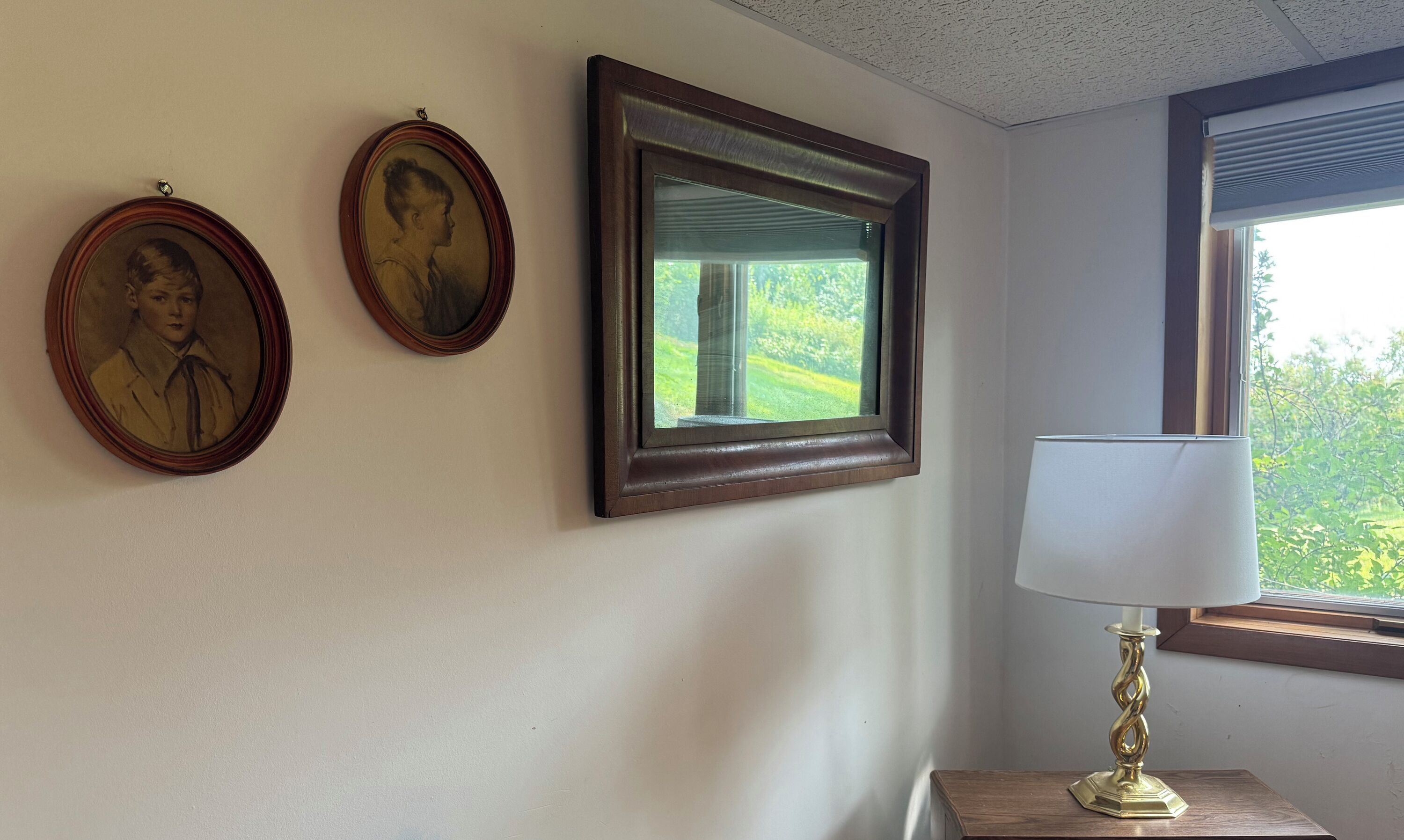 A room with three framed portraits on the wall, a window with a view of greenery, and a table with a lamp.