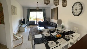 A modern living room with a dining area, a sofa, a TV, and a large wall clock. - Luxury complex 2 bed apartment with 5 pools and close to beaches, marina & golf (MANILVA)