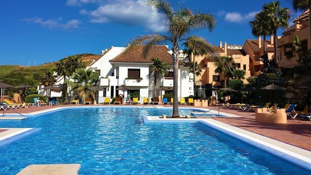 A swimming pool with lounge chairs and palm trees, surrounded by buildings with Mediterranean architecture. - Luxury complex 2 bed apartment with 5 pools and close to beaches, marina & golf (MANILVA)