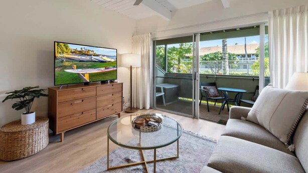 A living room with a TV, sofa, glass coffee table, and a balcony with outdoor furniture. - Remodeled Ground floor studio, AC, across from the beach, close to everything (Kihei)