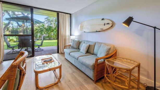 Una habitación con sofá, una mesa de centro de vidrio, sillas de mimbre, una tabla de surf en la pared y una lámpara.