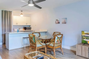 A modern kitchen with white cabinets, a dining area with wicker chairs, and a flat-screen TV.