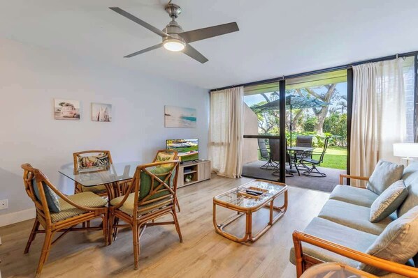 A living room with a glass coffee table, wicker chairs, a sofa, a ceiling fan, and a TV.