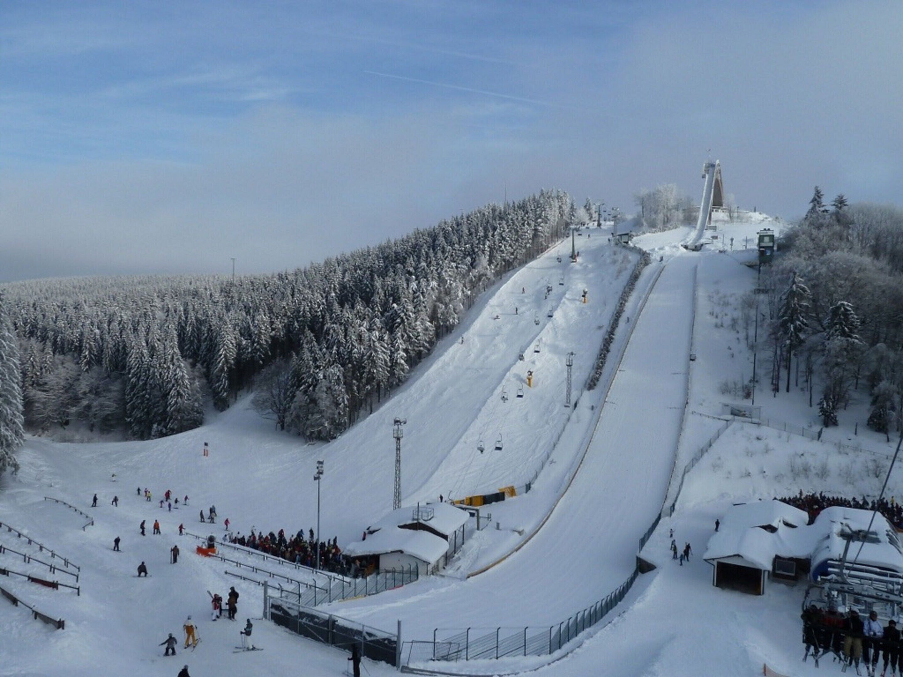 Ein mit Schnee bedeckter Skipisten mit Sessellift, bewaldetem Gebiet und einem kleinen Gebäude.
