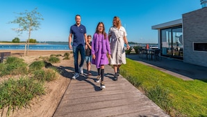 A family walking on a wooden boardwalk by a body of water, with a modern building and outdoor seating area in the background. - Chalet on the Water at the Beautiful Veluwemeer (Nunspeet)