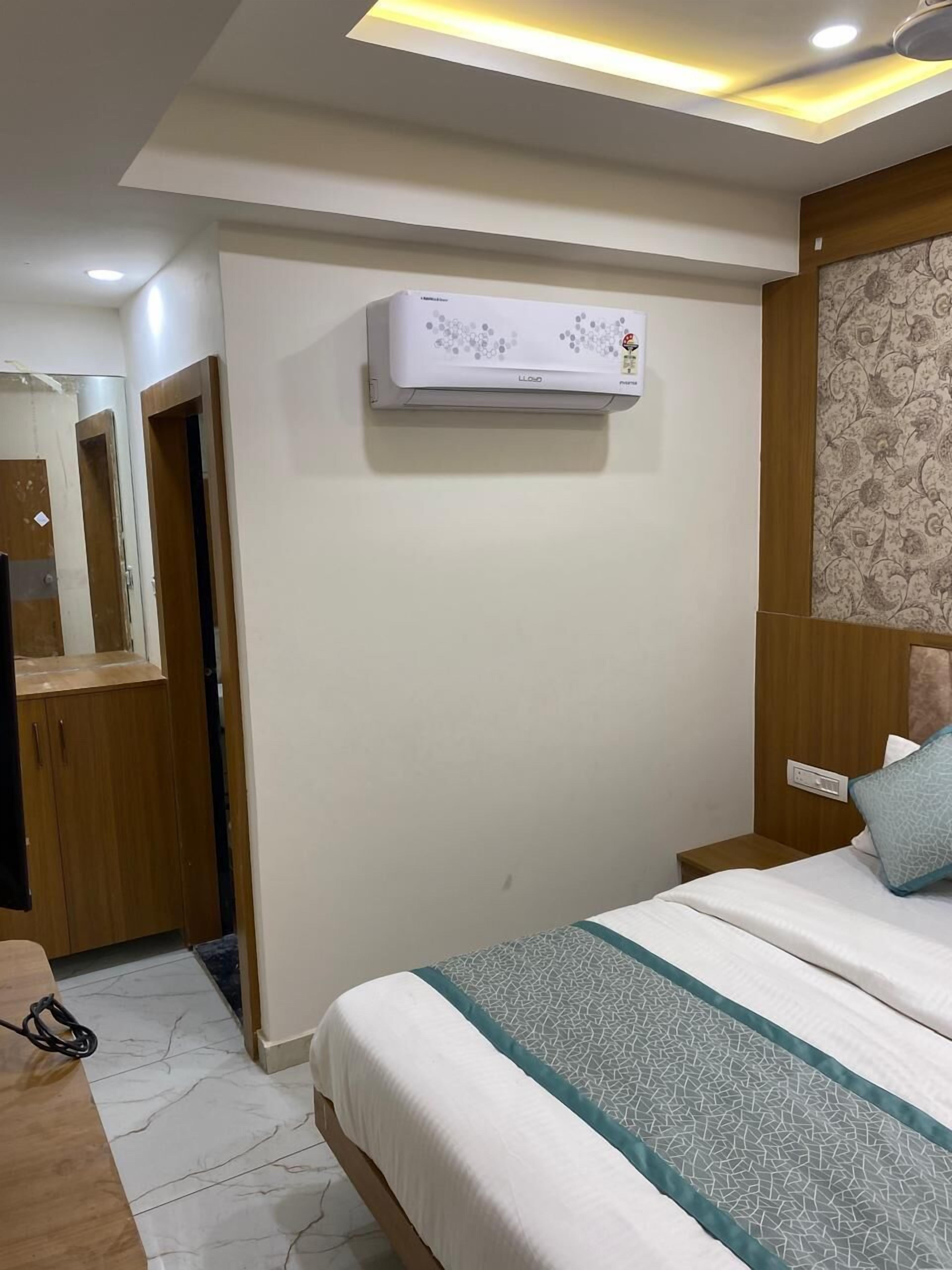 A hotel room with a bed, a wooden wardrobe, a mirror, an air conditioning unit, and a patterned wallpaper.