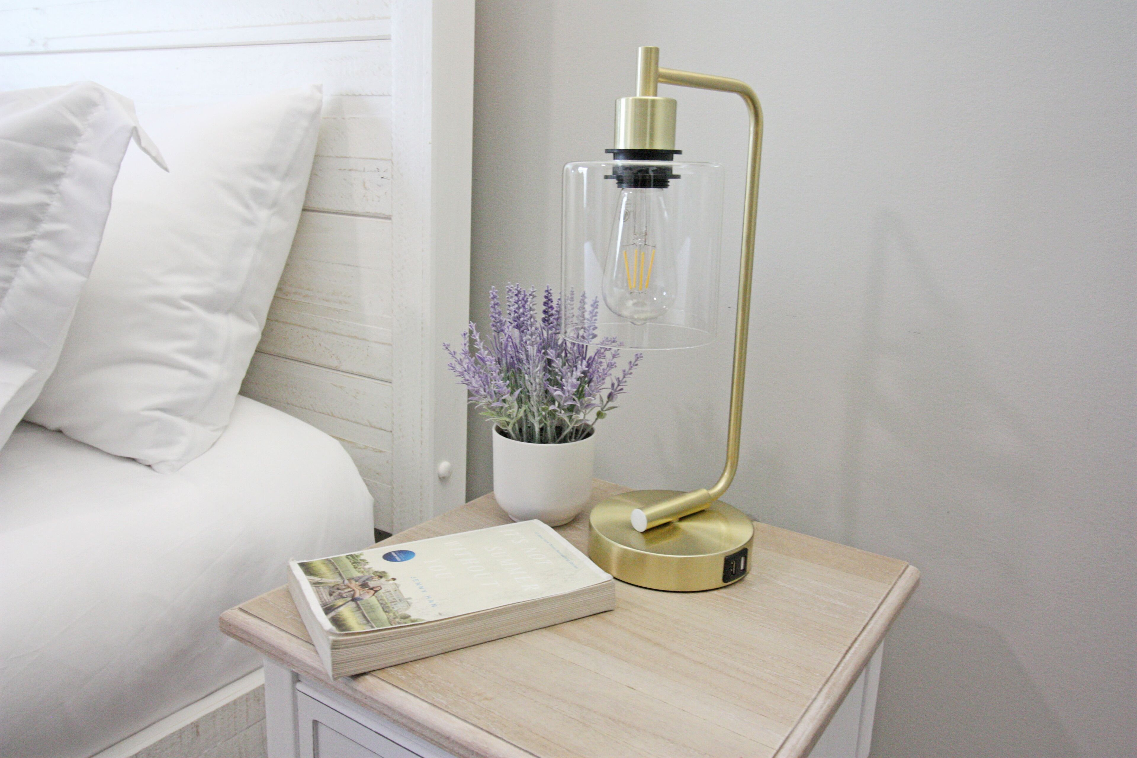 Un lit bien fait, avec une literie blanche, une table de chevet sur laquelle repose un livre, un pot de lavande et une lampe moderne dorée.