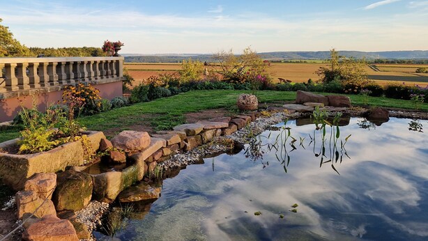 Een tuin met een vijver, stenen muren en uitzicht op velden en heuvels.