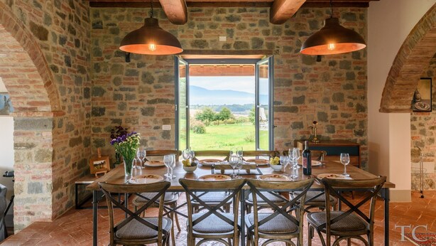 Un coin repas rustique avec un mur en pierre, des poutres en bois au plafond et une table dressée pour six personnes, avec des chaises.
