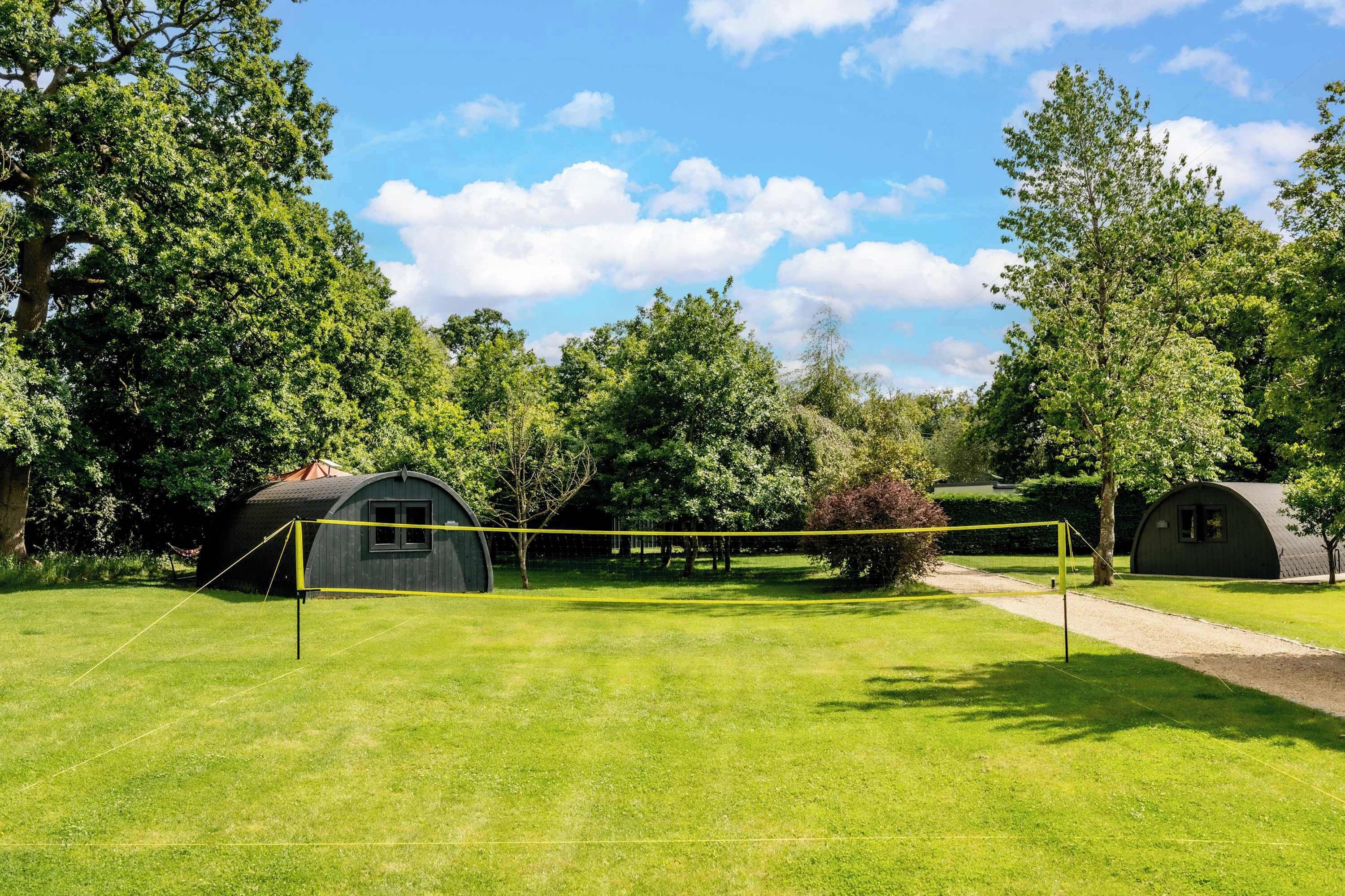A grassy field with a volleyball net, surrounded by trees and two small buildings.
