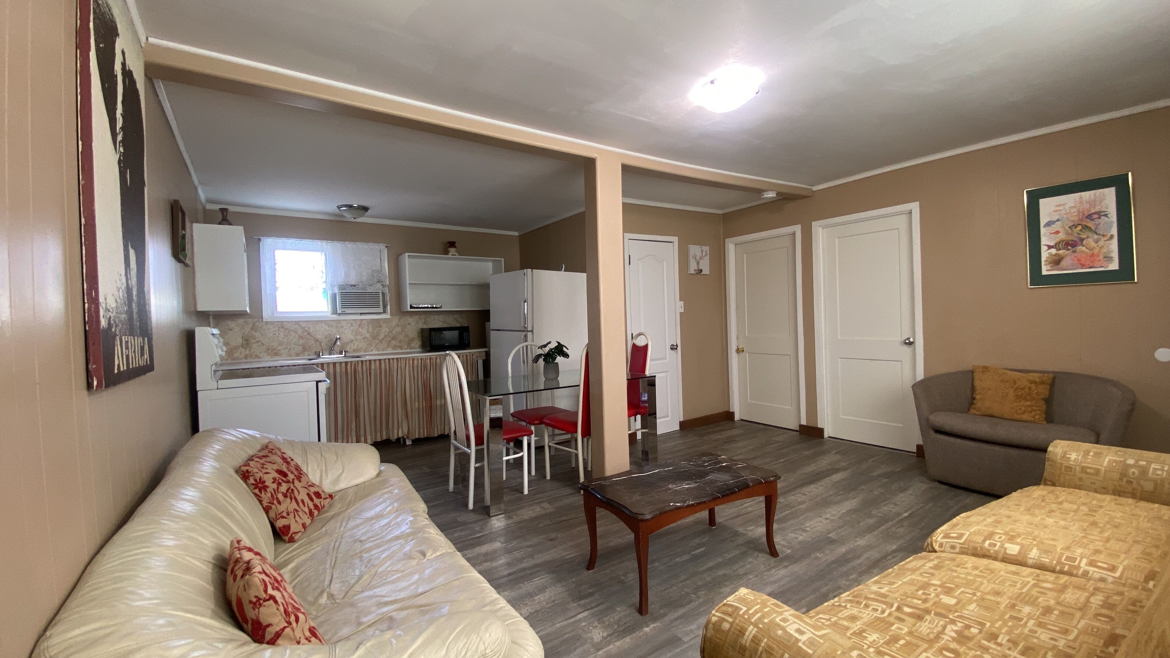 A living room with a sofa, a coffee table, and a couch. There is a kitchen area with a refrigerator, microwave, and sink. The room has a door and a window.