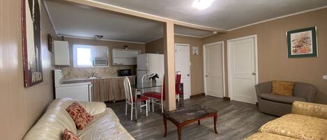 A living room with a sofa, a coffee table, and a couch. There is a kitchen area with a refrigerator, microwave, and sink. The room has a door and a window.