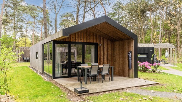 Eine moderne Holz-Blockhütte mit Glastür, einer Terrasse mit Tisch und Stühlen und Blick auf den umliegenden Wald.