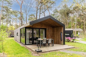A modern wooden cabin with a glass door entrance, a patio with a table and chairs, and a view of the surrounding forest. - Chalet in Eindhoven Near Bospark 't Wolfsven (Mierlo)