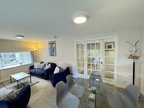 A modern living room with a glass dining table, dark sofas, and a large window with a view.