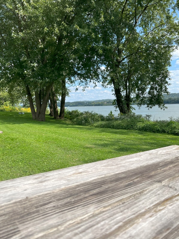 A wooden deck overlooking a grassy area with trees and a lake. - Cozy 2-bedroom RV on the Ohio River near historic Madison, IN with AC & a view! (Madison)