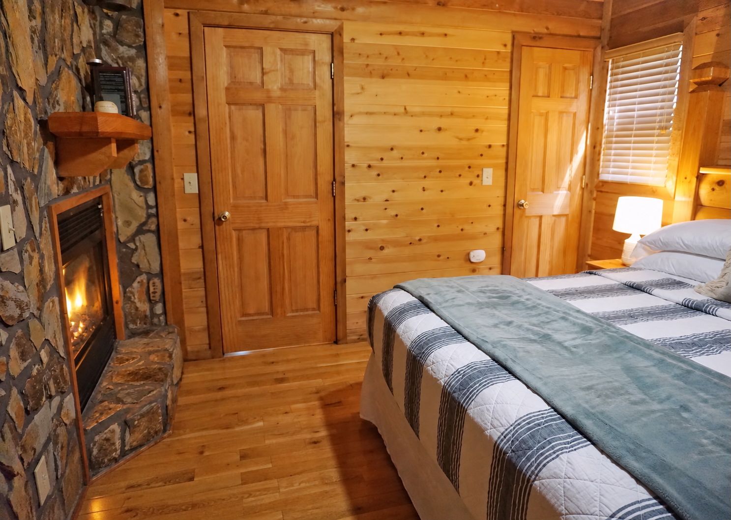 Une pièce chaleureuse avec une cheminée en pierre, des murs en bois, un lit recouvert d’une couverture à rayures et une table de chevet avec une lampe.