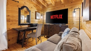 A cozy log cabin room with a wooden ceiling, a television displaying 'NETFLIX', a sofa, a desk with a chair, and a mirror.