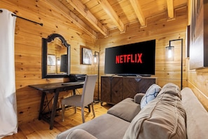 Une pièce chaleureuse aménagée dans un chalet en rondins, avec un plafond en bois, un téléviseur diffusant du contenu « NETFLIX », un canapé, un bureau avec une chaise et un miroir.